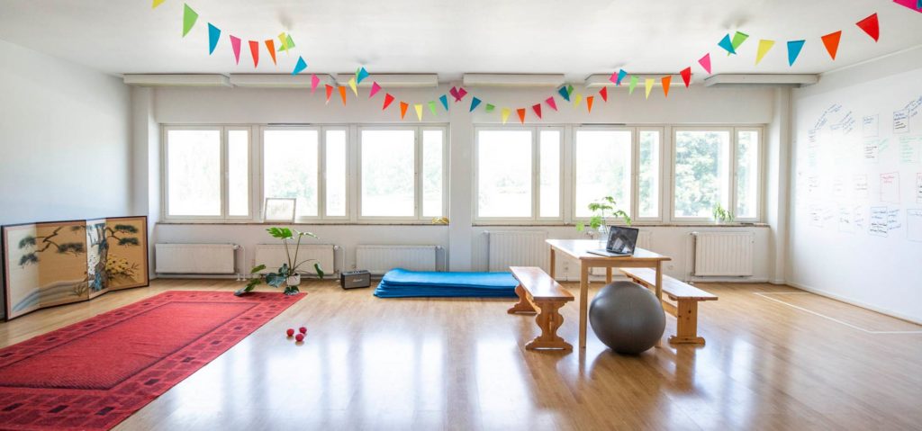Kuvassa valoisa tanssisali, jossa katossa värikkäitä lippuja, vasemmalla punainen matto ja oikealla pirtinpöytä ja penkit. Pöydän vieressä harmaa jumppapallo.