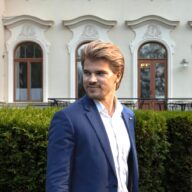 Mies sinisessä puvussa katsoo ohi kameran, taustalla kartano ja puska.
