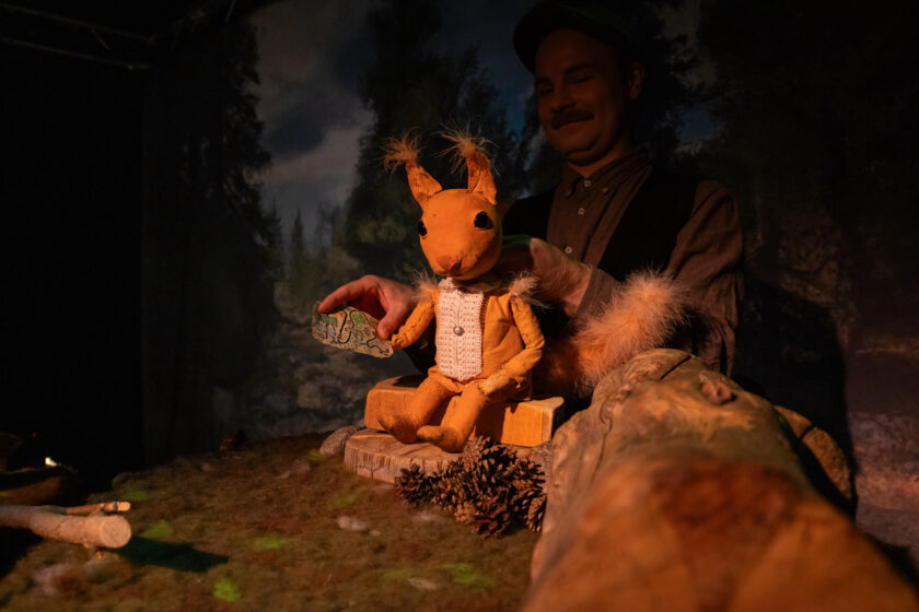 Kuvassa on kaunis metsäinen lavaste. Keskellä kuvaa istuu oranssi Orava-nukke ja hänellä on pieni aarrekartta kädessään. Taustalla pimeässä näkyy nukkea ohjaava nukettaja eli näyttelijä.