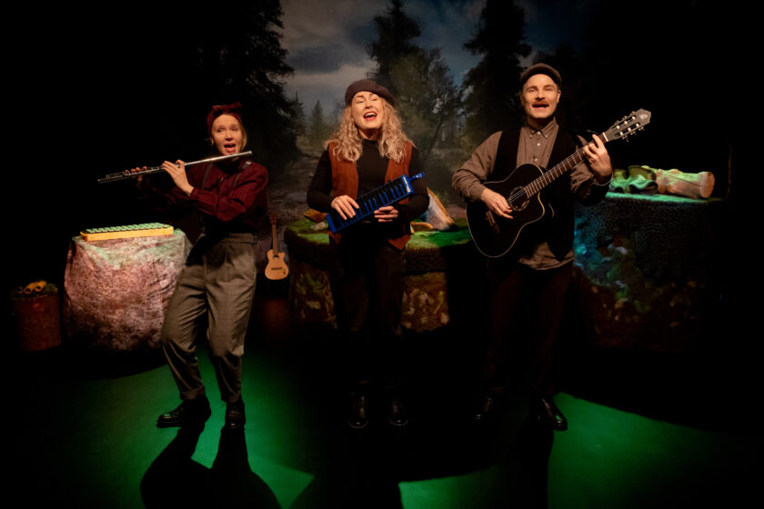 Kuvassa on näyttämön metsäisessä tunnelmassa musisoiva iloinen kolmikko. Vasemmassa reunassa oleva nainen soittaa huilua ja laulaa. Keskellä oleva nainen laulaa ja hänellä on kädessään tummansininen melodika. Oikealla hymyilevä, hattupäinen mies soittaa akustista kitaraa. Värimaailma on luonnon läheinen ja lattia on valaistu vihreäksi.