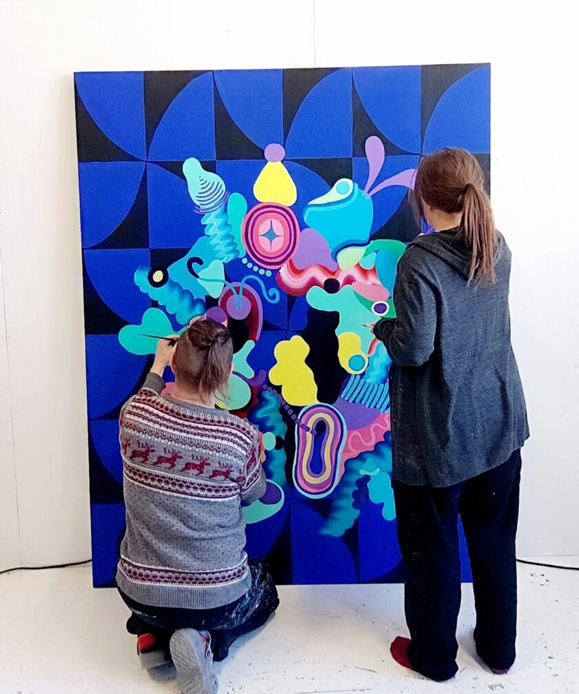 Inka Hannula ja Teemu Raudaskoski maalaamassa isoa värikästä abstraktia yhteismaalausta. 190 x 150 cm kokonen maalaus nojaa seinää vasten ja taiteilijat maalaavat vierekkäin.