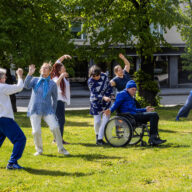 Kuvassa on aikuisia ihmisiä tanssimassa ulkona nurmikolla kesällä. Yksi on pyörätuolissa.