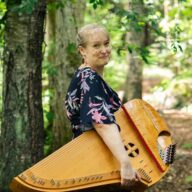 Kuvassa nainen seisoo metsäpolulla konserttikantele sylissä.