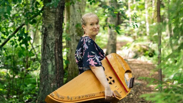 Kuvassa nainen seisoo metsäpolulla konserttikantele sylissä.