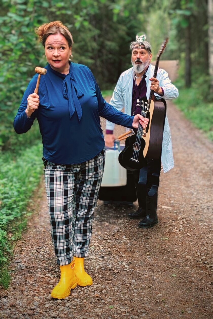 Nainen ja mies kävelevät polulla, naisella kädessään puheenjohtajan vasara ja ukulele. Miehellä lääkärin varusteet ja kitara kädessä.