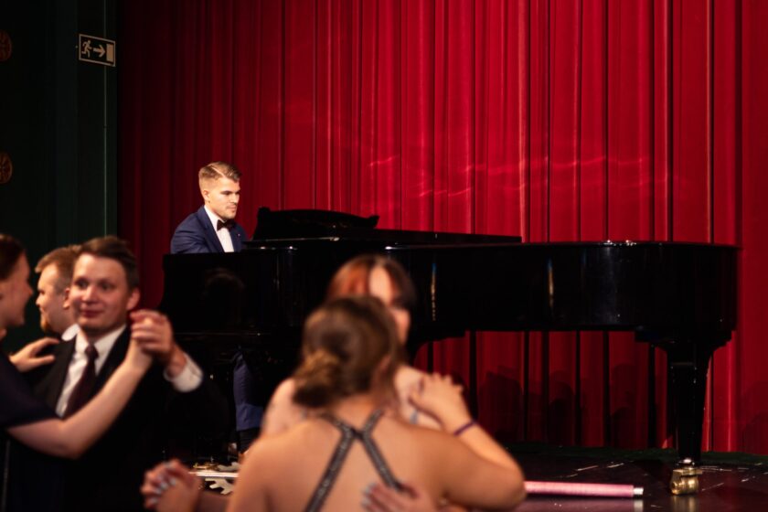 Nuori mies esiintyy flyygelin ääressä punaisen teatteriverhon edessä. Hän on pukeutunut tummansiniseen pukuun, valkoiseen paitaan ja ruskeaan rusettiin. Esiintymistilanne on keskittynyt ja arvokas, ja lämmin valo korostaa soittajan kasvoja sekä verhojen kangaspintaa. Kuvan etualalla tanssii juhlavasti pukeutuneita juhlavieraita.