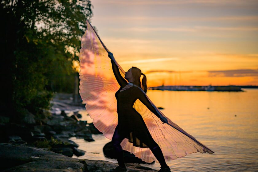 Aurongonlasku rannalla. Esiintyjä ojentaa käsiä jolloin siipien kangas kuultaa kauniisti auringon laskua vasten.
