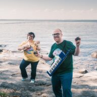 Kuvassa muusikot Paloma Catalá Parga ja Iago Castro Pérez seisovat kallioisella rannalla. Parga seisoo taka-alalla ja soittaa ukulelea pilli suussaan. Etualalla Pérez soittaa marakasseja, ja hänen kaulassaan roikkuu kosketinsoitin olkahihnassa.