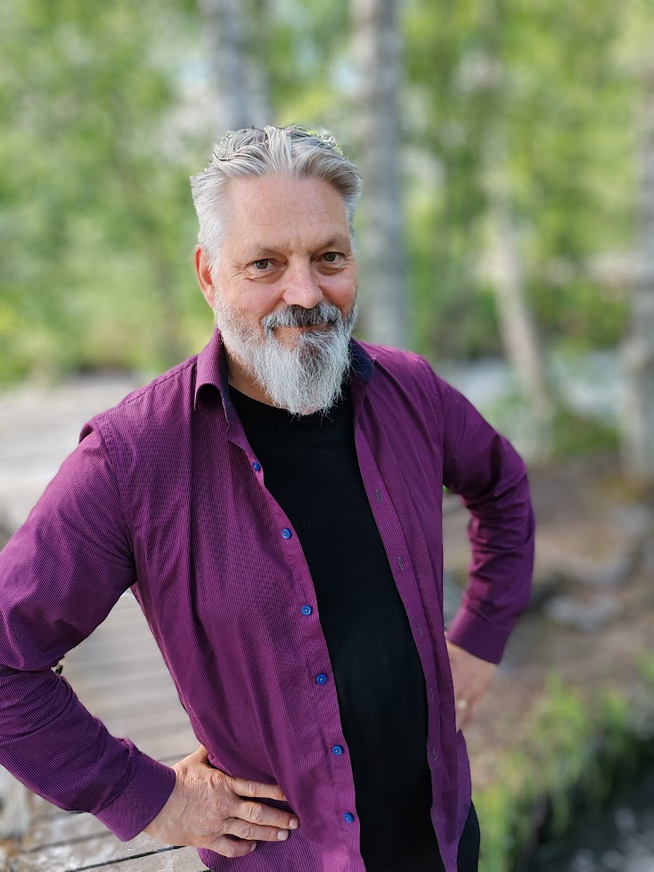 Kuvassa etualalla parrakas henkilö violetissa paidassa, taustalla epätarkasti puita.