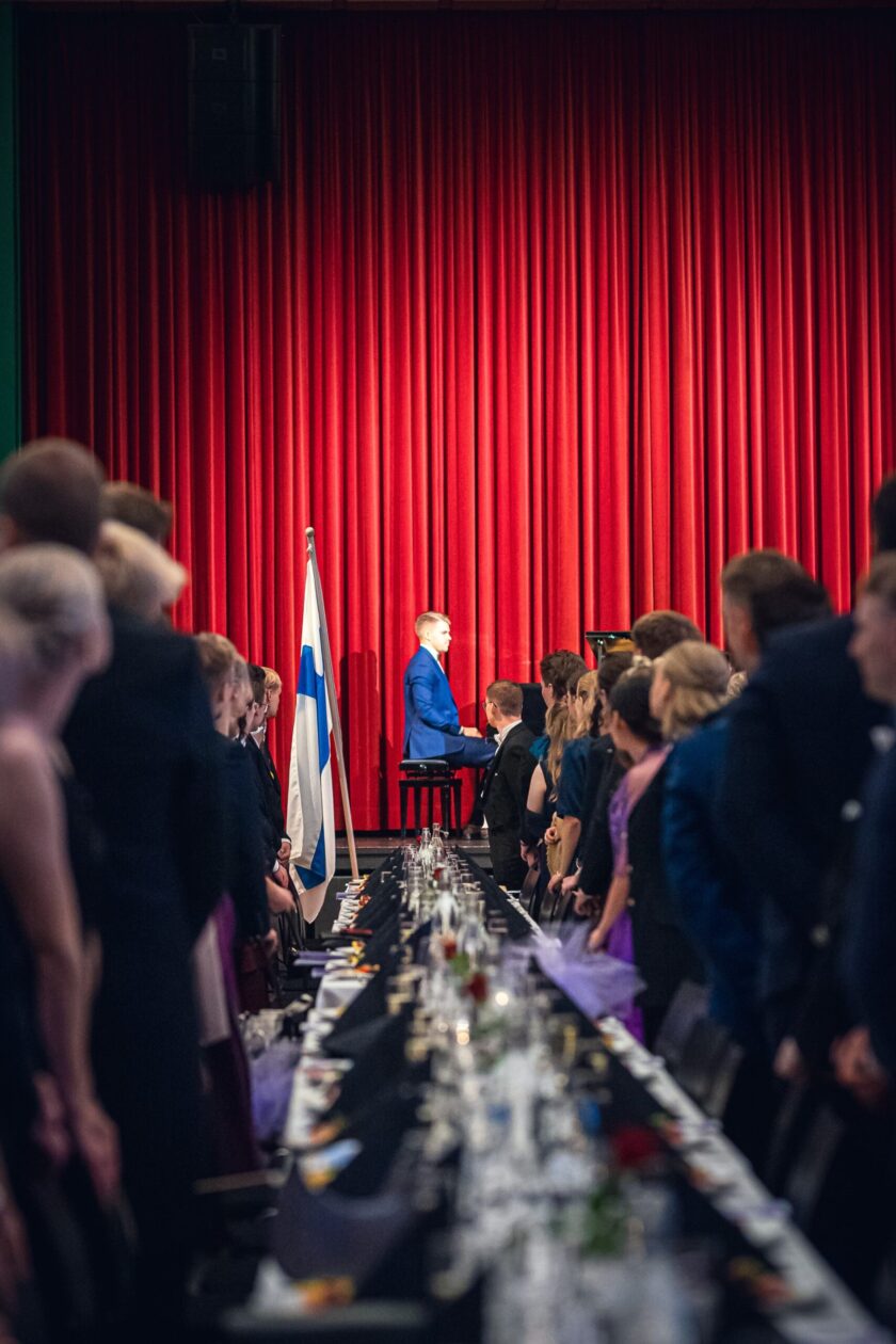 Juhlava juhlasalikuva, jossa pitkän juhlapöydän molemmilla puolilla seisoo juhlavasti pukeutuneita ihmisiä. Kuvan keskellä, lavan edustalla kirkkaanpunaisen teatteriverhon edessä, istuu siniseen pukuun pukeutunut nuori mies pianon ääressä esiintymisvalmiina. Pianistin vieressä seisoo Suomen lippu. Katsojat katsovat kohti pianoa seisten, tunnelma on arvokas ja odottava.