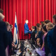 Juhlava juhlasalikuva, jossa pitkän, mustaliinaisen juhlapöydän molemmilla puolilla seisoo juhlavasti pukeutuneita ihmisiä. Kuvan keskellä, lavan edustalla kirkkaanpunaisen teatteriverhon edessä, istuu siniseen pukuun pukeutunut nuori mies pianon ääressä esiintymisvalmiina. Pianistin vieressä seisoo Suomen lippu. Katsojat katsovat kohti pianoa seisten, tunnelma on arvokas ja odottava.