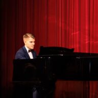 Nuori mies esiintyy flyygelin ääressä punaisen teatteriverhon edessä. Hän on pukeutunut tummansiniseen pukuun, valkoiseen paitaan ja ruskeaan rusettiin. Esiintymistilanne on keskittynyt ja arvokas, ja lämmin valo korostaa soittajan kasvoja sekä verhojen kangaspintaa.