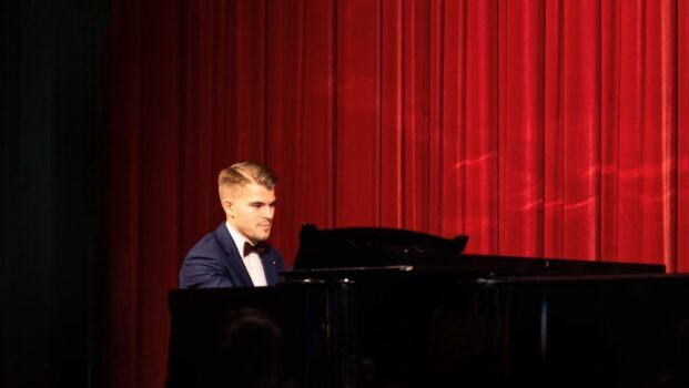 Nuori mies esiintyy flyygelin ääressä punaisen teatteriverhon edessä. Hän on pukeutunut tummansiniseen pukuun, valkoiseen paitaan ja ruskeaan rusettiin. Esiintymistilanne on keskittynyt ja arvokas, ja lämmin valo korostaa soittajan kasvoja sekä verhojen kangaspintaa.