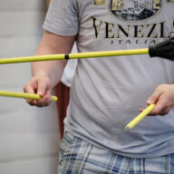 Kuvassa on kaksi kättä, joissa on kaksi keltaista keppiä ja ilmassa on yksi keltainen keppi, jongleerausvälineenä flower stick, jonka henkilö on heittänyt.